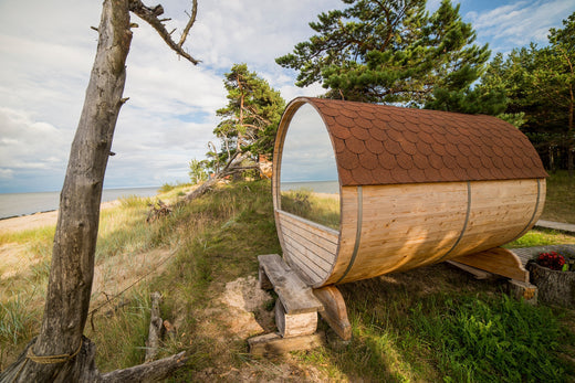 Houten buitensauna op een zand- en grasrijke omgeving bij een strand, met uitzicht op de zee en omgeven door bomen en vegetatie
