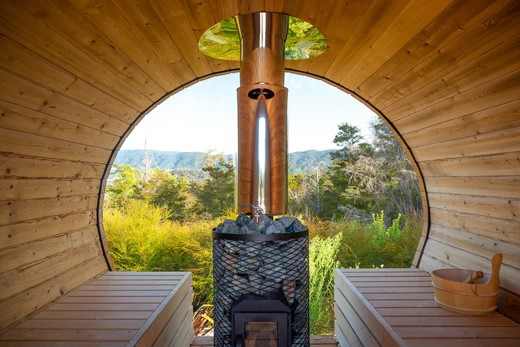 Interieur van een houten buitensauna met twee banken, een houtkachel en uitzicht op een natuurlijke omgeving door een groot rond raam