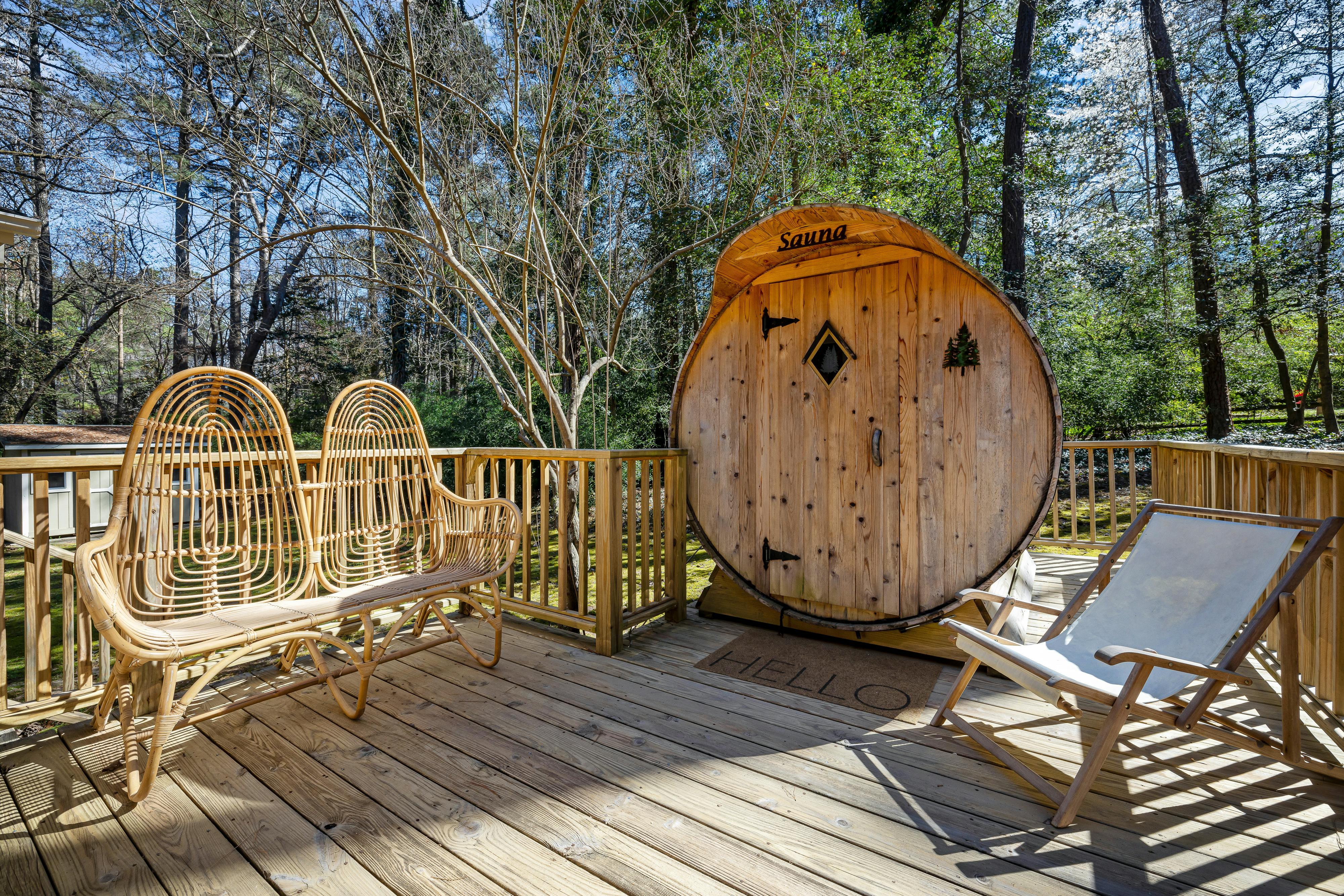 Ronde houten sauna met een deur en raam, geplaatst op een houten terras met rieten stoelen en ligstoelen, omringd door bomen